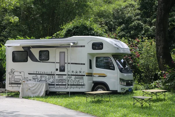Yeşil parka park edilmiş beyaz bir eğlence aracı var. Üzerinde evler ve çocukların çizimleri var. Yanında bereketli ağaçlarla çevrili bir açık hava masası var..