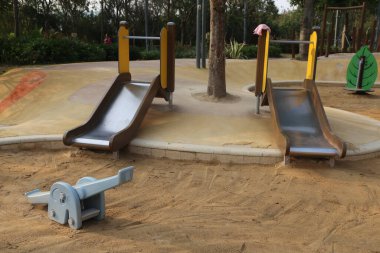 Kumlu bir çocuk parkı, iki slayt ve metalden yapılmış bir tırmanma iskeleti. Arka planda, ağaçları ve koltuk veya bank gibi diğer park ekipmanlarını görebilirsiniz.