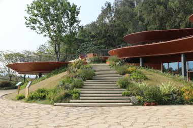 Kıvrımlı, kırmızı toprak bina bir tepenin üzerinde, yemyeşil ve ağaçlarla çevrili. Dış duvarlarda soyut bir manzara yaratan paslı çelik paneller var.