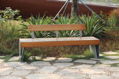 Kenarlarında paslanmaz çelik bacakları olan ahşap ve metalden yapılmış basit bir park bankı yeşil bitkilerle çevrili bir açık alana yerleştirilmiş.