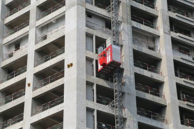 boş bir binanın yan tarafında asılı kırmızı iskele asansörü, gri beton duvarlar ve zeminler, her penceresinde çelik ızgara olan sütunlar.