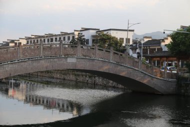 Taştan bir kemer köprüsü, yolun her iki tarafında beyaz duvarlar ve siyah fayanslar bulunan antik Çin mimarisi tarzında nehri kaplar. Köprünün yanında, iki sıra cadde boyunca diğer binalarla çevrili eski bir ev duruyor..