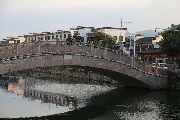 Taştan bir kemer köprüsü, yolun her iki tarafında beyaz duvarlar ve siyah fayanslar bulunan antik Çin mimarisi tarzında nehri kaplar. Köprünün yanında, iki sıra cadde boyunca diğer binalarla çevrili eski bir ev duruyor..