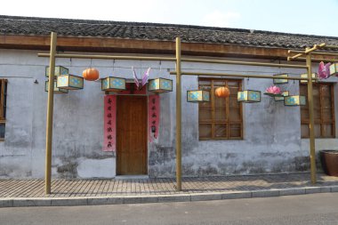 Çin kırsalındaki eski bir evin beyaz duvarı fenerler ve ahşap sütunlarda asılı renkli afişlerle süslenmiş. Bir uçtaki küçük bir kapı iç tarafa açılıyor..