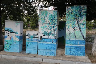 Kentsel bir yolun kenarında, üç buzdolabı şekilli nesne titiz mavi ve yeşil manzara resimleriyle boyandı. Bu aletlerdeki dolapların şekilleri ve renkleri farklıydı..
