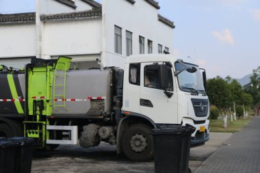 Küçük çöp kamyonu beyaz binanın önüne park edilmiş, her iki tarafında yeşil çöp tenekeleri ve gri çimento kaldırımları var. Kamera tüm aracı bir açıdan yakalıyor, görüntü detaylarını gösteriyor..