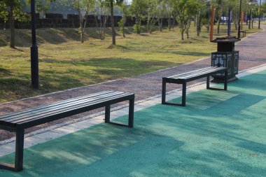 Asfalt yolun sol tarafında yeşil çimenler, ağaçlar ve çiçekler olan bir parka. Yolda oturulacak siyah metal sokak mobilyaları var.