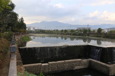 Net bir gökyüzü, uzak dağlar, binalar ve arkasında görünen yollar ile bir kentsel alandaki su ve kara bloğu manzarası
