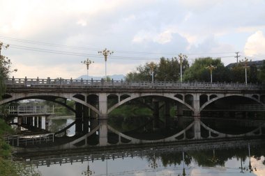 Nehir üzerinde bir köprü: eski bir taş kemer köprüsünün dört yarı dairesel kıvrımlı serbest fotoğrafı, yüzeyine yansıyor, açık bir gökyüzü ve bulutların altında.