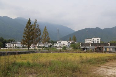 Uzakta, Çin kırsalındaki pirinç tarlaları ve ağaçlarla çevrili terk edilmiş beyaz bir bina ve arkasında dağlar var..