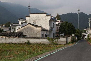 Güney Çin 'in kırsal kesimlerinde, yanında siyah kiremitleri ve gri duvarları olan eski beyaz duvarlı bir ev var. Şu eski binanın önünde iki bina daha var.