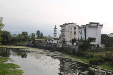Uzakta, beyaz duvarları ve siyah kiremitleri olan eski bir bina var. Bu binaların önünde bir kat daha gri taş ev duruyor.