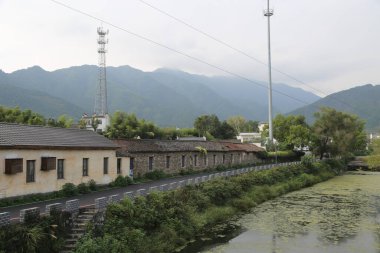 Uzakta, Çin 'in kırsal kesimlerinde yeşil bitki örtüsüyle çevrili alçak duvarları ve kiremit çatıları olan iki evin bir tarafında eski bir bina var..