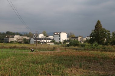 Uzakta, Çin kırsalında eski beyaz bir bina var. Yanında birkaç küçük ev ve yakınında pirinç tarlaları var..