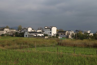 Beyaz binalar, yeşil alanlar ve arka planda gri bir gökyüzü olan bir köy. Önünde insanların sebze ektiği açık bir alan var..