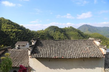 Yeşil dağlar ve mavi gökyüzü ile çevrili eski Çin çiftlik evinin çatı kiremitleri görünürde hiç bulut yok. Birkaç evin beyaz duvarları her iki taraftaki yemyeşil ağaçların arka planına karşı duruyor.