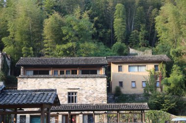 Güney Çin 'in dağlık bölgesinde, tuğla ve taş dış duvarları ve fayanslı çatıları olan iki modern evin resmi. Üstü yeşil ağaçlarla örtülmüş.