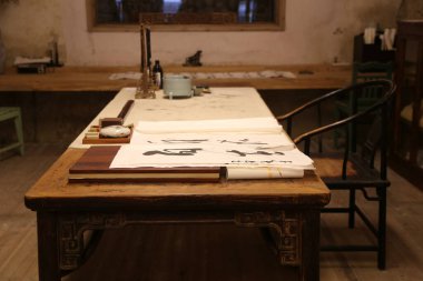 Kaligrafi sınıfında üzerinde iki büyük beyaz kağıt çarşaf olan eski bir masa var. Karakterlerin ön görünümü ve mürekkepli fırça darbeleri yakın çekim görüntülerinde görülebilir.