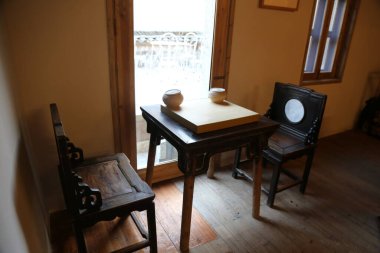 Eski bir Çin evi çalışma odasında bir ahşap masa ve iki sandalye var. Ayrıca her sandalyenin önünde iki tane beyaz kare taş var. Duvarlarda açık sarı boya var.