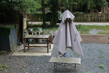 Gri bir pelerin açık bir masanın üzerine örtülmüş, arkası bize bakıyor. Sahne, evin önündeki yemyeşil ve ahşap çitlere karşı gölgesini yakalıyor.