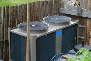 İrlanda 'da bir evin çatısına yerleştirilmiş ve arkasında bambu çitleri olan hava kaynaklı bir ısı pompasının detaylı fotoğrafı. Birim gri ve mavi, en üstte iki büyük dairesel pervane var..