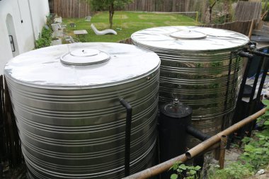 İki büyük paslanmaz çelik su tankı, üzerinde beyaz kapaklar olan bir şehir bahçesinin arka bahçesinde. Soldaki tank, yanındaki tanktan daha uzun ve daha küçük..