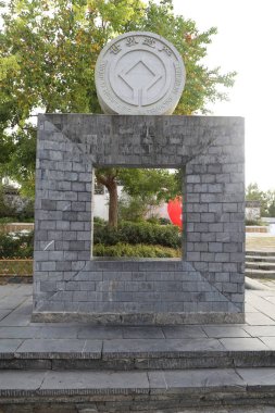 Üzerinde gri tuğlalar ve kare ve dikdörtgen şeklinde beton bloklar bulunan, açık havada ağaçların altında bulunan, üzerinde oymalı ulusal amblem olan kare taştan bir anıt..