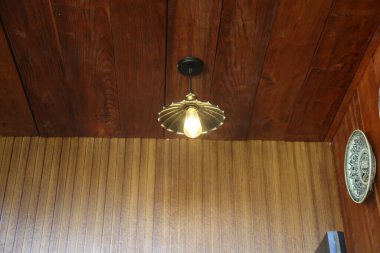 Tavandan sarkan, ahşap panelin üstünde asılı antik bir pirinç lamba armatürü. Ampul yanar ve hafifçe parlar. Etrafında başka mobilya ya ya da dekorasyon parçası yok..