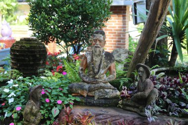 Tayland 'da, Chiang Mai' deki eski bir bungalovun bahçesinde çapraz bacaklı bir keşişin taş heykeli var. Yanında maymun benzeri evcil iblisiyle meditasyon yapıyor..