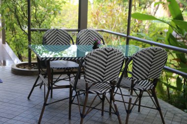 Sembol desenli siyah beyaz rattan sandalyeler, açık verandada cam tepeli yemek masası. Açık hava balkonunun köşesinde, tropik bir ortamda bahçeye bakan siyah bir hasır sandalye.