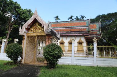Beyaz duvarları ve altın aksanlı yeşil çimenler ve ağaçlarla çevrili küçük bir Tayland tapınağı. Kapı altın metalden yapılmıştır ve karmaşık tasarımları vardır..