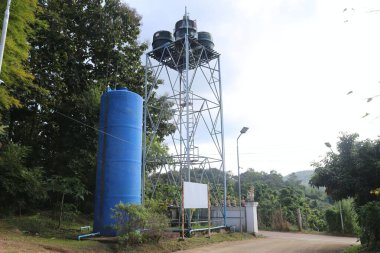 Tayland 'ın kırsal kesimindeki su tankı kulesi mavi ve yanında beyaz tabelalı bir elektrik jeneratörü var. Etrafta ağaçlar da olmalı..