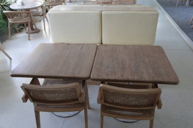 İki kare ahşap masa ve rattan sandalyeler, iki dikdörtgen koltuklardan biri beyaz ve modern bir restoranın açık hava alanında, beyaz beton zemini olan dört koltuğu var..