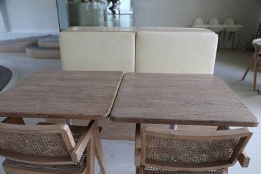 Önünde sandalyeler olan iki dikdörtgen ahşap masa. Yerde duruyorlar ve tahtada bazı çizikler var. Arka planda boş beyaz bir kanepe ve camdan bir sehpa var.