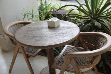 Yuvarlak ahşap bir masa, balkonunda iki tane rattan sandalye ve önünde de boş bir mendil kutusu. Arka planda yeşil bitkiler ve beyaz duvarlar var. Yeni ve doğal bir tarz yaratıyorlar..