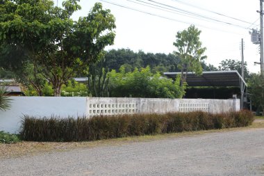 Evin önünde beyaz beton bir çit, yol kenarında, duvarın arkasında ağaçlar ve çalılar, sağ tarafta Tayland tarzı bir garaj.