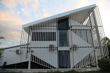 Dış duvarlarda iç içe geçmiş bir desen oluşturan, geometrik şekiller yaratan ve tasarımına görsel ilgi ekleyen açısal metal levhalarla beyaz bir bina.