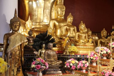 Tapınakta çeşitli çiçek ve süslemelerle çevrili bir grup altın Buda heykeli. Ana figür, elleri önünde katlanmış, zarif bir tahtta oturuyor.
