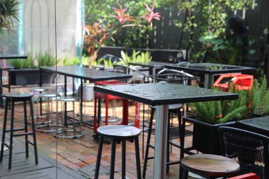 Kırmızı çelik bacaklı, yemyeşil bitkilerle çevrili siyah metal yüksek bar masalarının yer aldığı modern bir açık hava kafe. Masalar ve sandalyeler yağmurdan ıslanmış, ve ahşap döşemeler yağmur damlalarıyla kaplı.