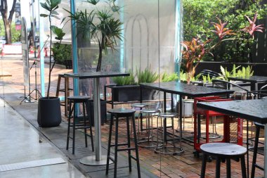 Öğleden sonra boyunca Jakarta kaldırımında saksılarla çevrili, modern yüksek masaları ve siyah metal bar tabureleri olan bir açık hava kafesi. Ayar, alan hissi veren cam duvarları içerirken, yakındaki binalar kentsel atmosp oluşturur.