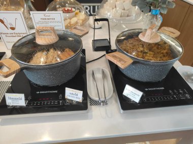 İki tencerenin üzeri siyah taş desenli ve cam kapaklı, bir tencere kahverengi pirincin yarısı, ortadaki elektrikli elektrikli ocak tarafından doldurulmuş, süpermarketin iç mekanının kahvaltı büfesi alanında.