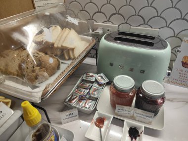 Bir tost makinesi, bir elektrikli ızgara ve beyaz tezgahın üstündeki küçük plastik kaplarda reçel. Önünde, içinde tek bir parça ekmek olan saydam bir kutu daha var.