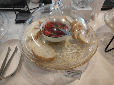 Bir ofis masasının önündeki iki açık kristal buz piramidinin üzerinde duran şeffaf bir tabağın içine konmuş domates bruschetta ile dolu bir kase ve dilimlenmiş cam bir saat.