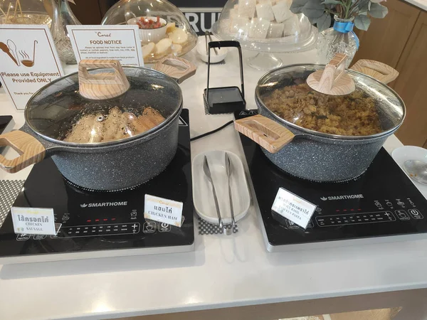 İki tencerenin üzeri siyah taş desenli ve cam kapaklı, bir tencere kahverengi pirincin yarısı, ortadaki elektrikli elektrikli ocak tarafından doldurulmuş, süpermarketin iç mekanının kahvaltı büfesi alanında.