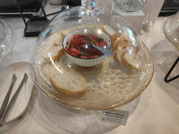 Bir ofis masasının önündeki iki açık kristal buz piramidinin üzerinde duran şeffaf bir tabağın içine konmuş domates bruschetta ile dolu bir kase ve dilimlenmiş cam bir saat.