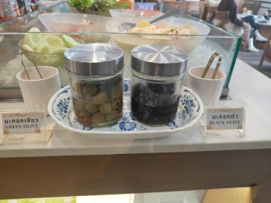 İçinde siyah zeytin ve yeşil zeytin bulunan iki cam kavanoz havaalanı kafesinin ortasındaki zarif beyaz bir tabağın üstüne yerleştirilmiş.