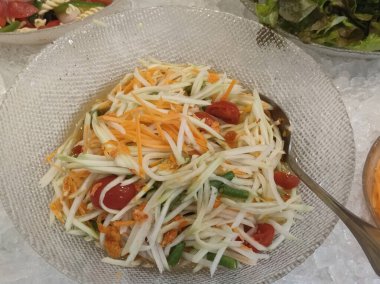 Büyük bir yığın yeşil papaya salatası, som tam, doğranmış havuç ve salatalık beyaz tabağın sağ tarafında buz gibi yuvarlak cam bir masada oturuyor. Ayrıca içinde kırmızı domates de var..