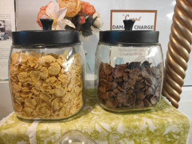 Bir sanat galerisinde ticari ürün fotoğrafçılığı için sergilenen masanın üstünde mısır cipsi ve çikolata kaplı pirinç pufları bulunan iki cam kavanoz.