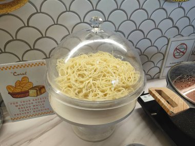 Şeffaf plastik pasta standı spagetti erişteleriyle dolu, mutfak tezgahına elektrikli buharlı tencerenin yanına yerleştiriliyor ve güvenliğiniz için iki karton iskambil kağıdı konuyor..