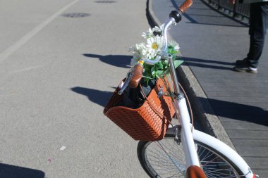 Önünde turuncu deri bir sepet olan beyaz bir bisiklet, içinde küçük bir buket çiçek ve bir çift siyah güneş gözlüğü var. Motosiklet kameraya doğru bir açıda duruyor.